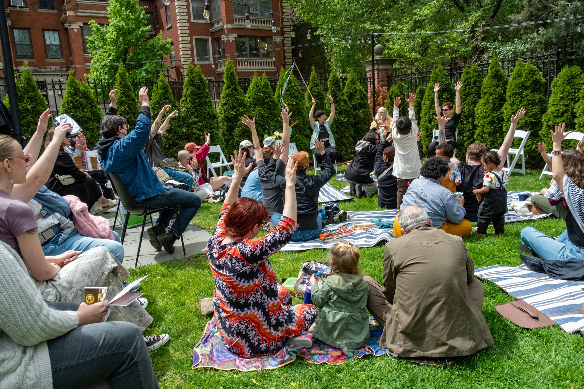 A group of people of all ages sit together on blankets and chairs outside; they are all raising their arms up.