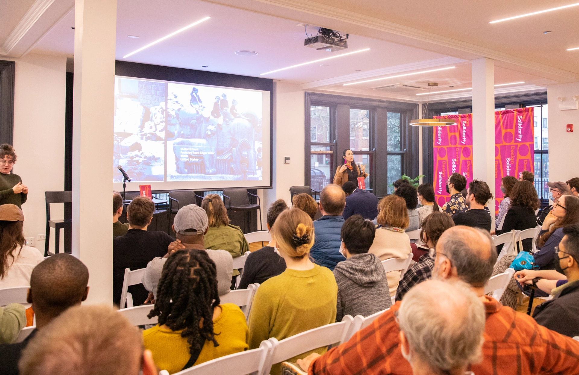 A large audience listens to a speaker, who is showing slides on a projected screen.